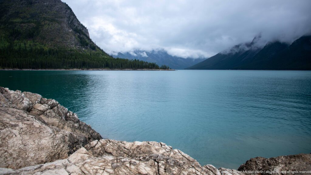 Wide Lake Minnewanka view from pale rocks toward forested mountains and a mist-filled valley.