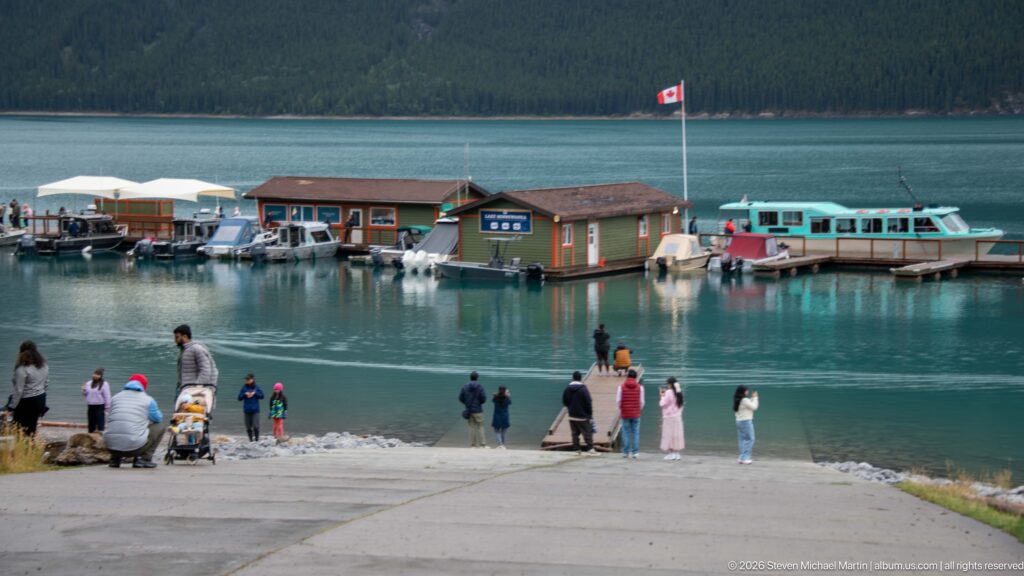 Visitors gathered at the Lake Minnewanka shoreline facing the marina, tour boats, Canadian flag, and forested opposite shore.