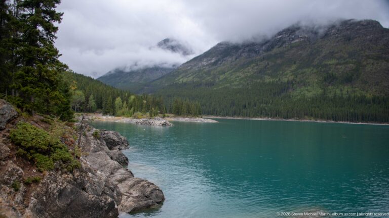 Rocky shore beside turquoise Lake Minnewanka with forested mountains disappearing into cloud.