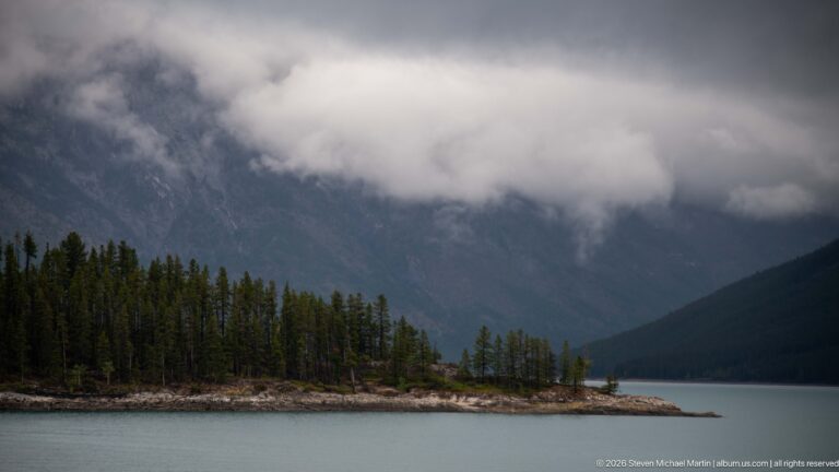 Forested point extending into Lake Minnewanka below a mountain obscured by low clouds.