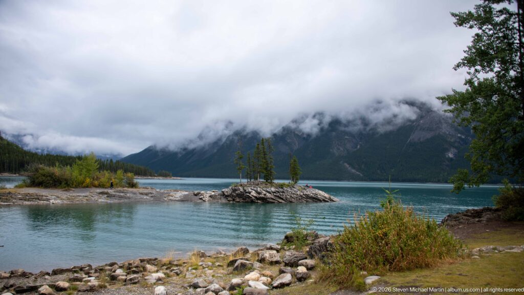 Rocky Lake Minnewanka shoreline looking toward small islets and cloud-covered mountains.