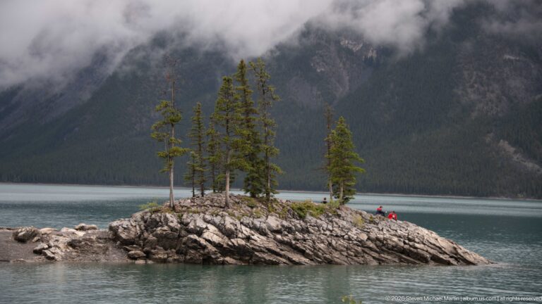 Small conifer-covered rocky islet in Lake Minnewanka with misty mountain slopes behind.