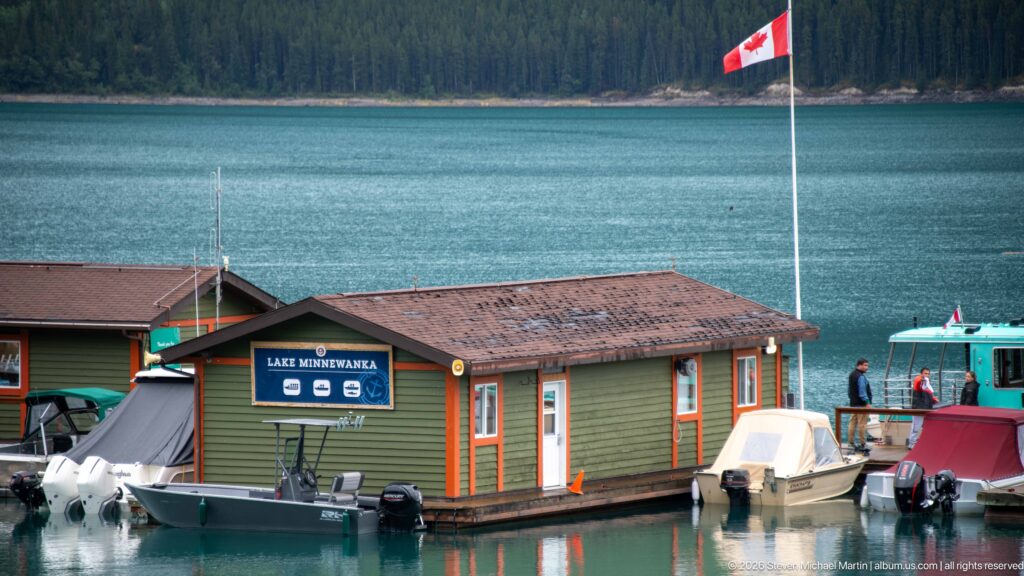 Marina building and small boats on turquoise Lake Minnewanka beneath a Canadian flag, with forest across the water.