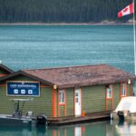 Marina building and small boats on turquoise Lake Minnewanka beneath a Canadian flag, with forest across the water.