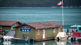 Marina building and small boats on turquoise Lake Minnewanka beneath a Canadian flag, with forest across the water.