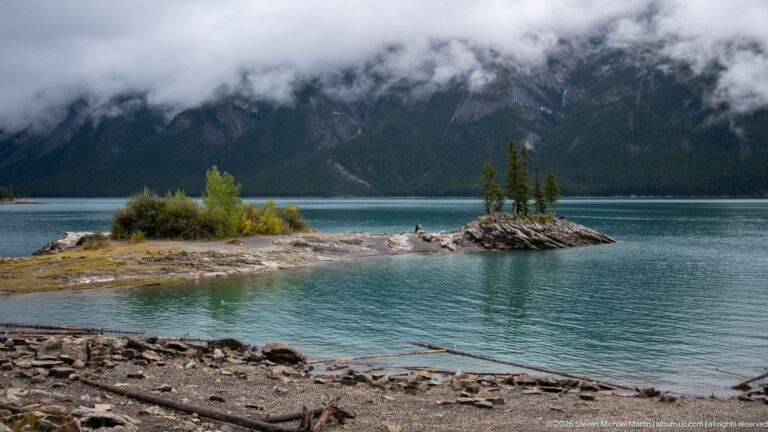 Small conifer-covered rocky islets in turquoise Lake Minnewanka below cloud-wrapped mountains.