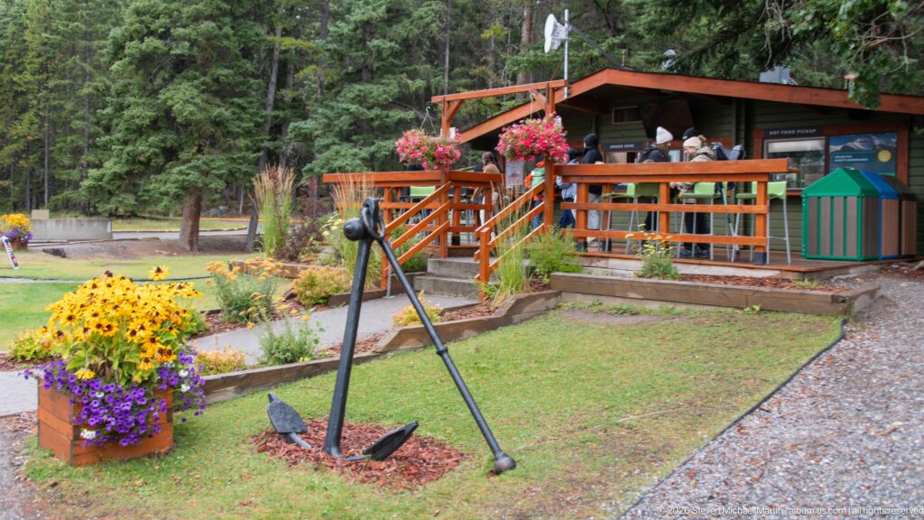 Flower planters and a large anchor stand before a rustic Lake Minnewanka service building.
