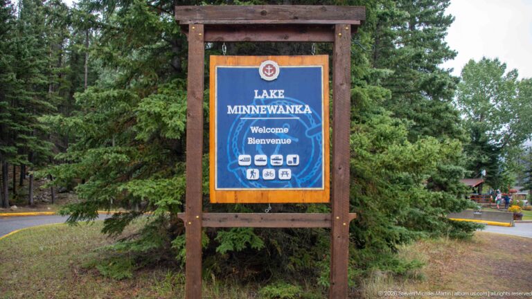 Timber-framed Lake Minnewanka welcome sign beside a conifer-lined access road.