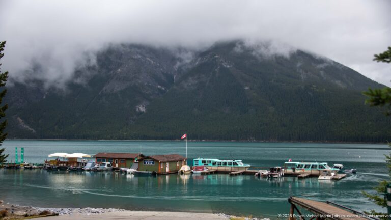 Lake Minnewanka marina and tour boats below a dark mountain slope partly hidden by low clouds.