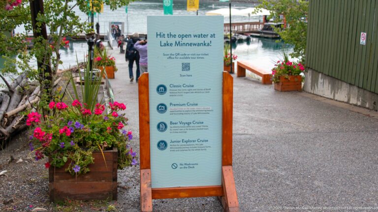 Cruise information sign beside flower planters on the walkway leading toward Lake Minnewanka marina docks.