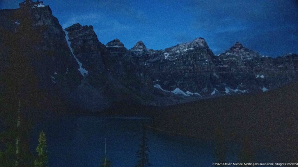 Dark twilight view of Moraine Lake beneath jagged mountain silhouettes and faint snowfields under a deep blue sky.