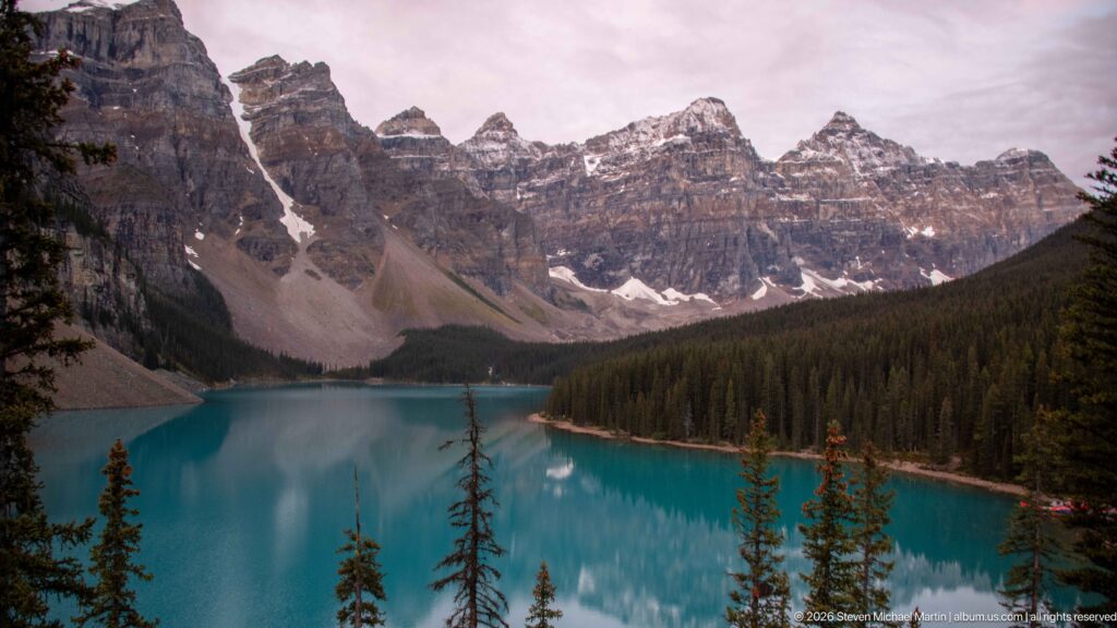 Elevated view of turquoise Moraine Lake, foreground conifers, forested shore, scree fans, snowfields, and jagged peaks.
