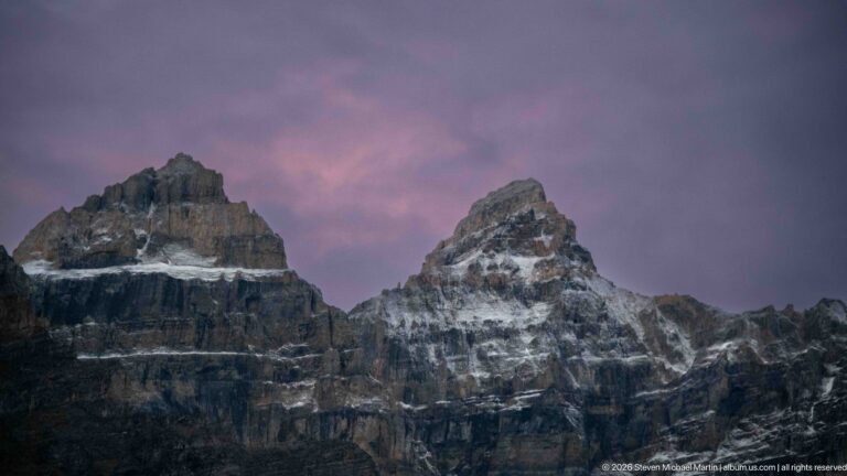 Two dark, snow-streaked Rocky Mountain summits silhouetted against a purple-pink evening sky.