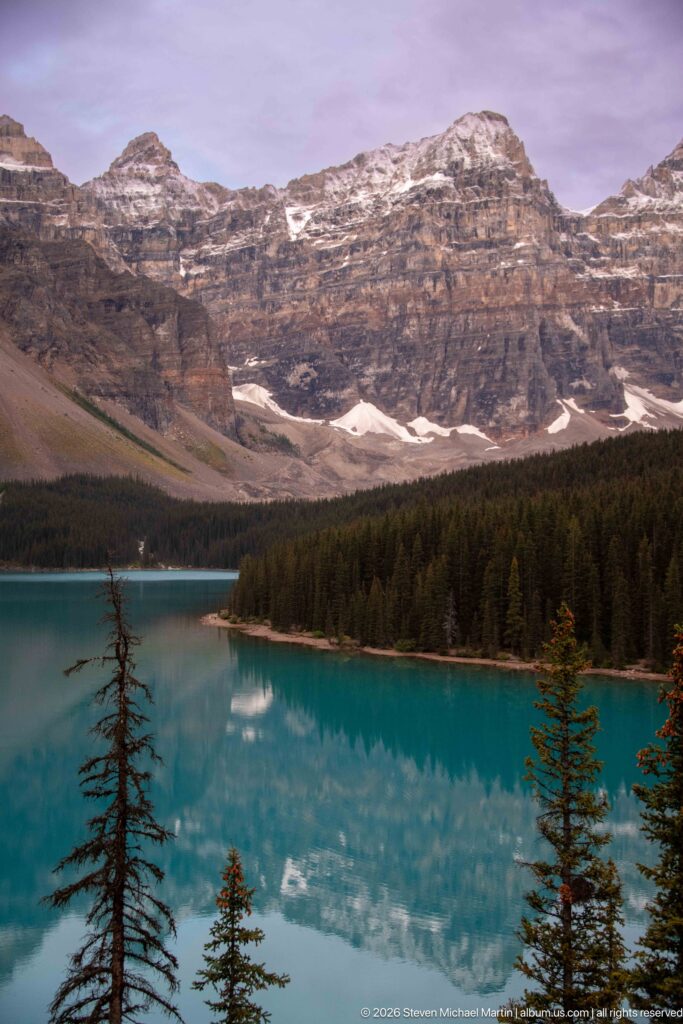 Turquoise Moraine Lake reflecting clouds below layered mountains, snow patches, forested shoreline, and foreground conifers.