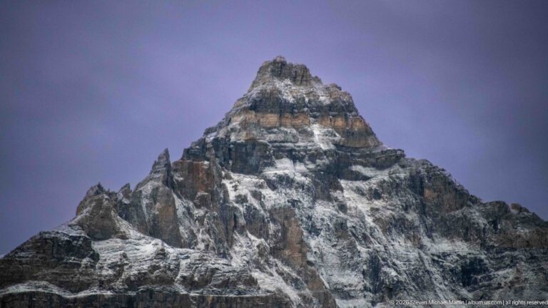 Close view of a steep layered mountain peak dusted with snow beneath a muted violet-gray sky.