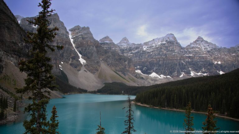 Moraine Lake stretching beneath jagged layered peaks, with turquoise water, conifer forest, scree slopes, and lingering snow.
