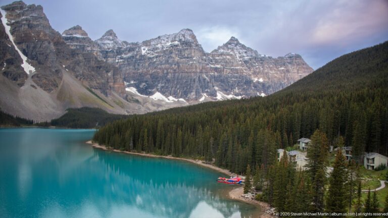 Turquoise Moraine Lake below rugged peaks, with dense conifer forest, red canoes, and lodge buildings along the shore.
