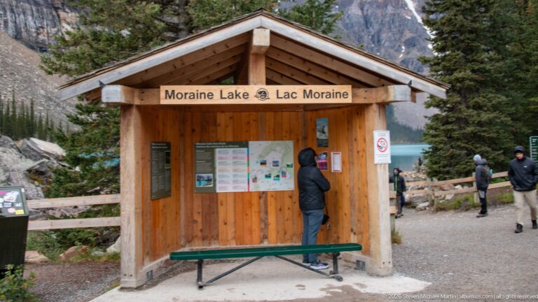 Wooden Moraine Lake information kiosk with bilingual sign, maps, bench, visitors, conifers, and turquoise lake beyond.