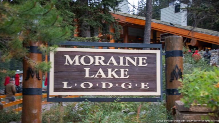 Close view of the Moraine Lake Lodge sign mounted between heavy timber posts, with lodge buildings and evergreens behind.
