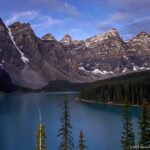 Moraine Lake in partial shadow beneath jagged peaks, with sunlit conifers, scree fans, snow patches, and blue sky.