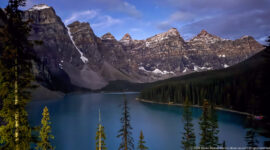 Moraine Lake in partial shadow beneath jagged peaks, with sunlit conifers, scree fans, snow patches, and blue sky.