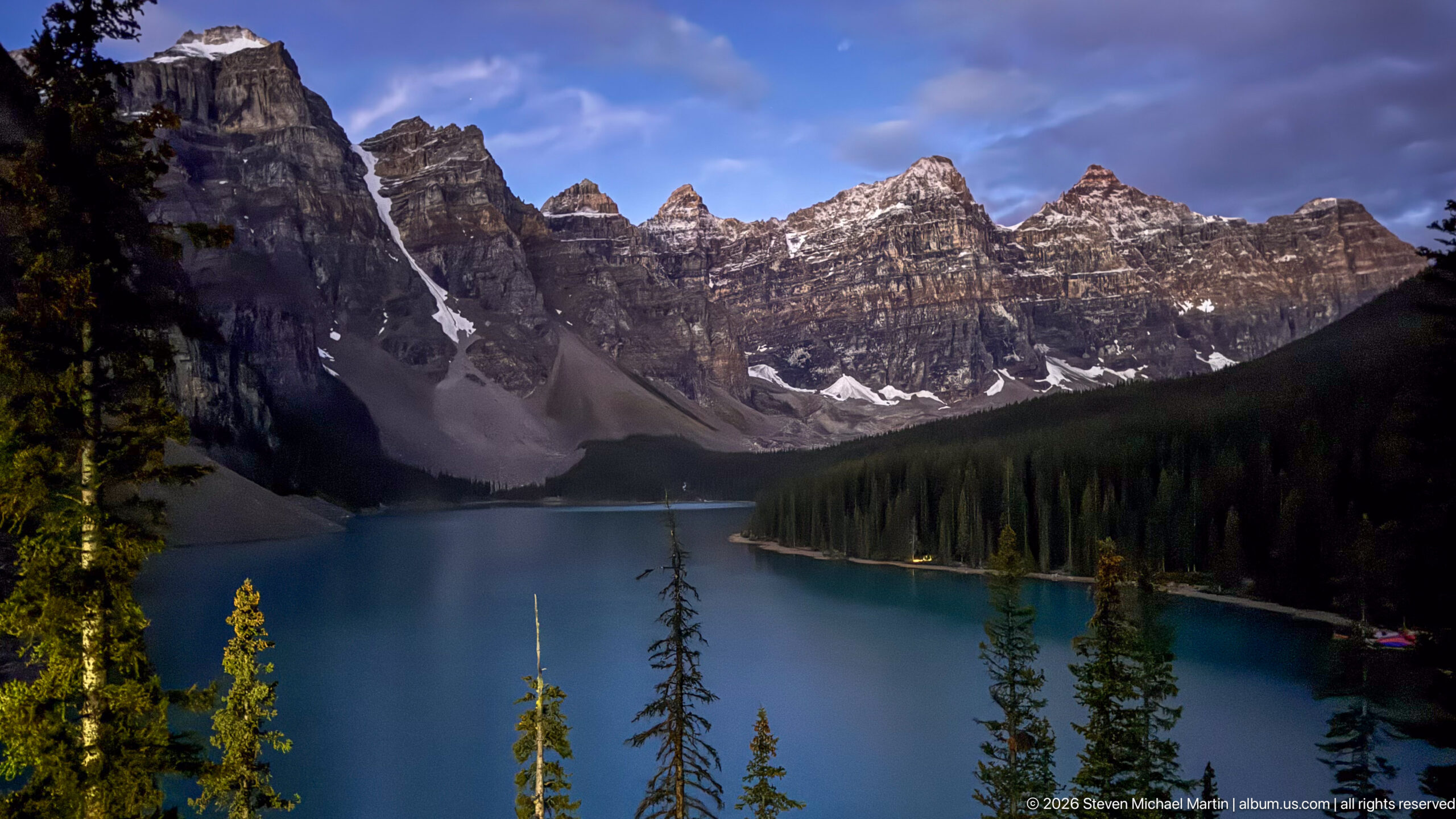 Moraine Lake in partial shadow beneath jagged peaks, with sunlit conifers, scree fans, snow patches, and blue sky.