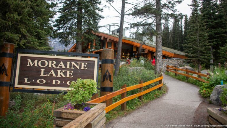 Moraine Lake Lodge sign beside a paved path, wooden railings, flowers, and lodge buildings among spruce trees.