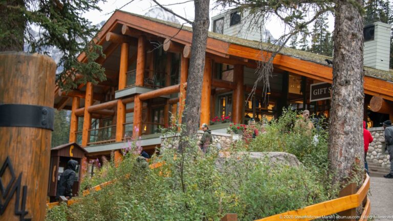 Large timber lodge with log posts, broad windows, stone base, conifers, wildflowers, and visitors near entrance.