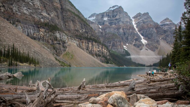 Driftwood and boulders beside turquoise Moraine Lake with rugged peaks, snow, forest, and visitors beyond.