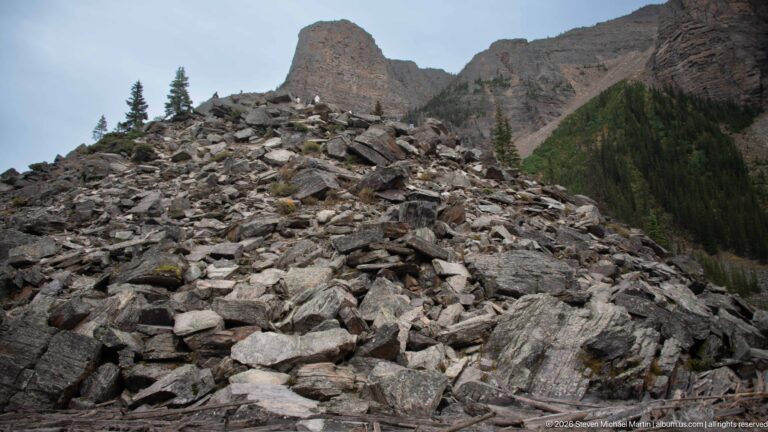 Close view across angular gray boulders on the Rockpile with sparse conifers and mountain cliffs behind.