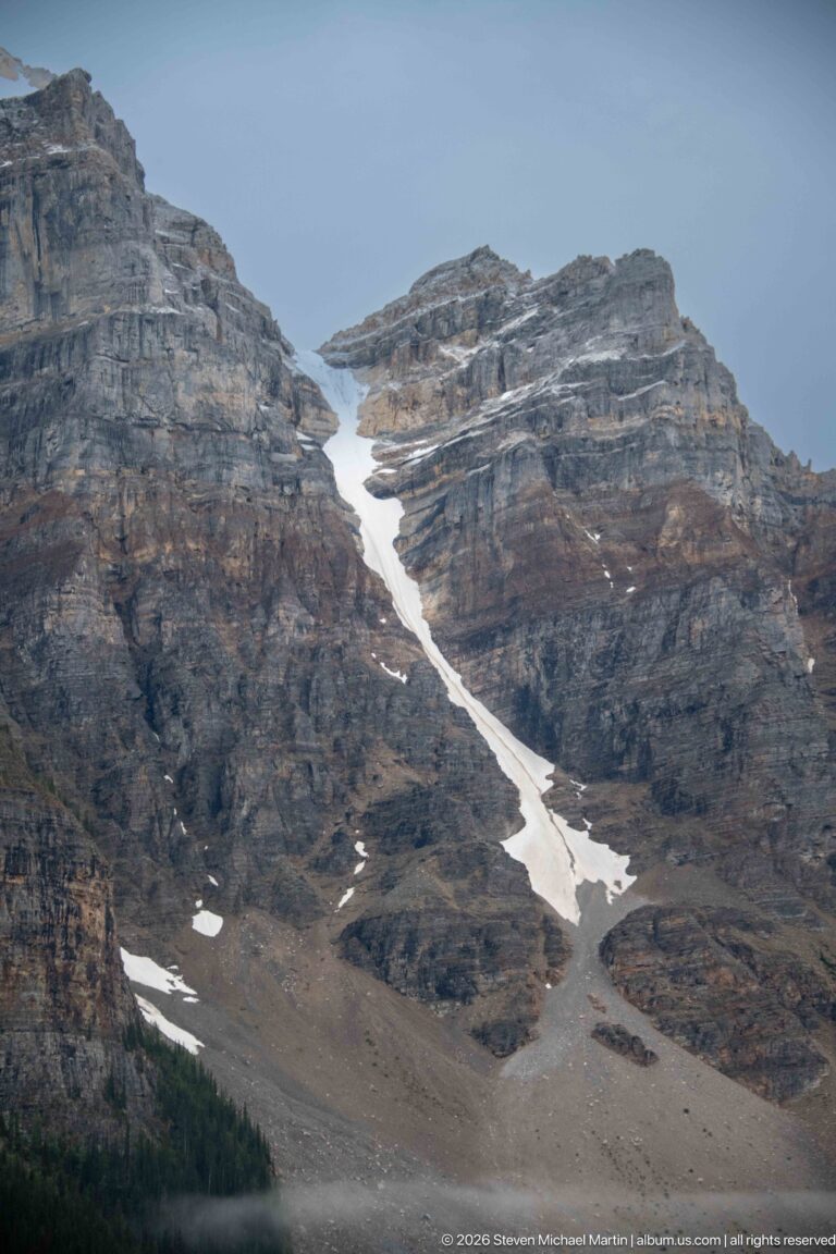 Narrow white snowfield descending through layered gray mountain cliffs and scree beneath an overcast sky.