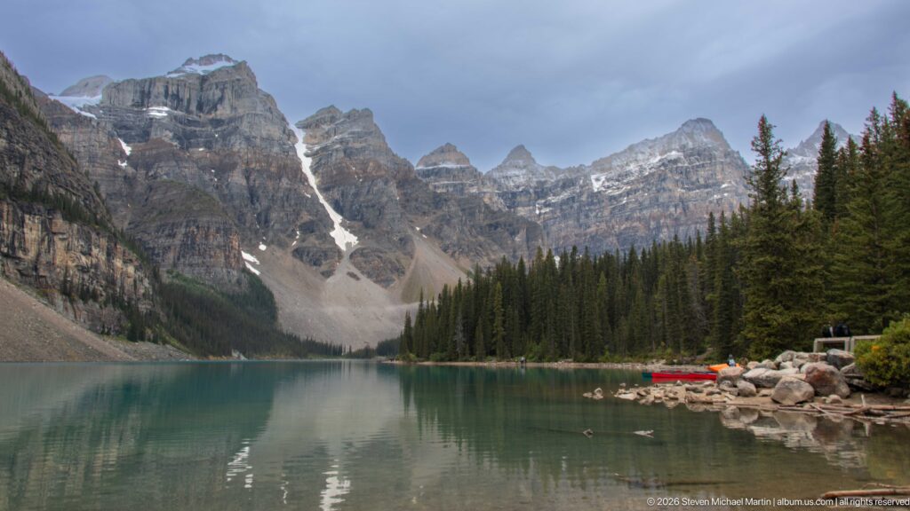Turquoise Moraine Lake beneath rugged gray peaks, snow-filled gullies, conifers, and a small red canoe.