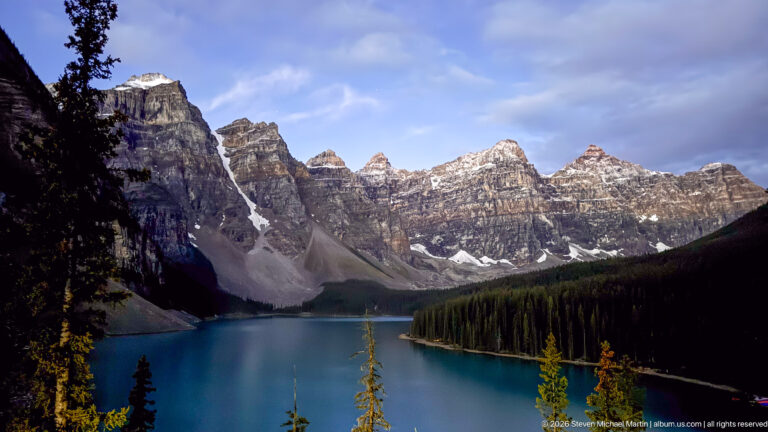 Moraine Lake beneath sunlit jagged peaks and clearing clouds, with dark forested shore, scree slopes, and foreground conifers.