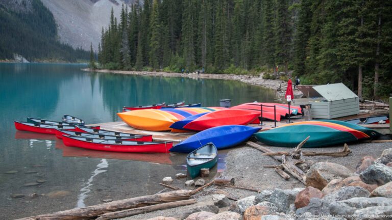 Rows of colorful canoes pulled onto the rocky turquoise shoreline beside dense evergreen forest.