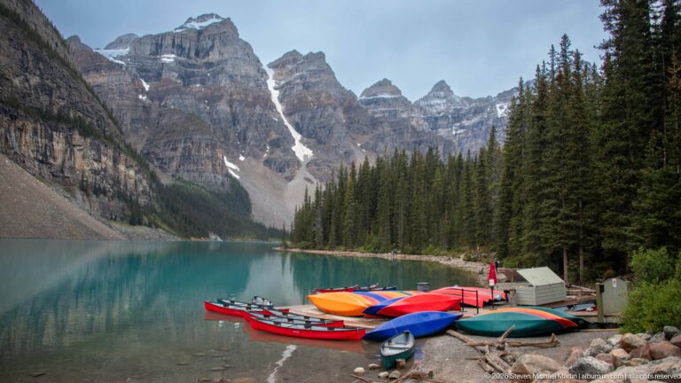 Colorful canoes on the turquoise shore of Moraine Lake beneath steep gray peaks and conifer forest.