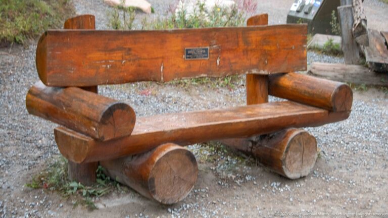 Rustic bench made from thick peeled logs with a small plaque on the wooden backrest.