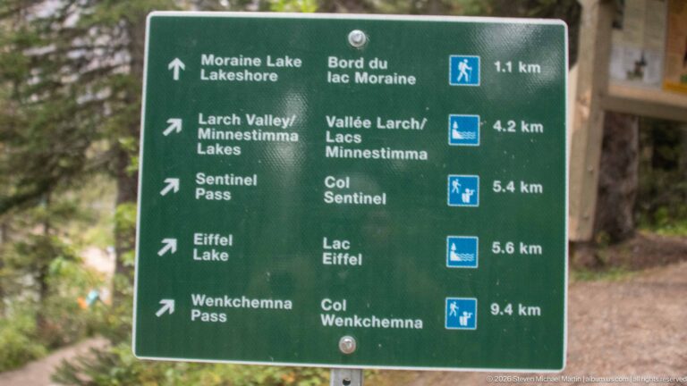 Green bilingual trail sign listing Moraine Lake Lakeshore, Larch Valley, Sentinel Pass, Eiffel Lake, and Wenkchemna Pass.