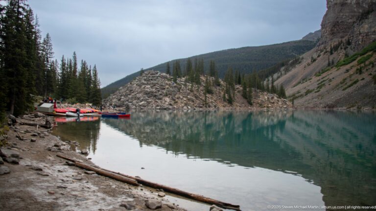Calm turquoise lake beside muddy shore, colorful canoes, conifers, and the boulder-covered Rockpile.