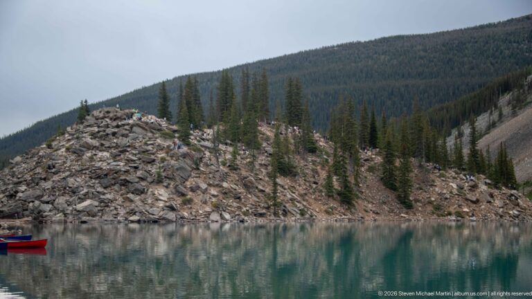 Rocky boulder mound with scattered conifers reflected in the turquoise water of Moraine Lake.
