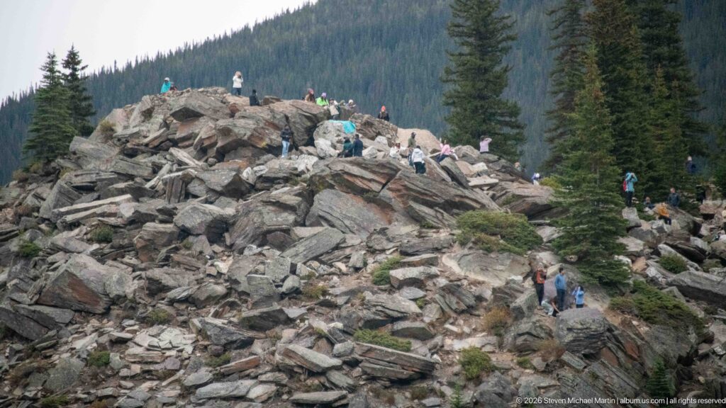 Visitors climbing a large mound of angular gray boulders dotted with conifers above Moraine Lake.