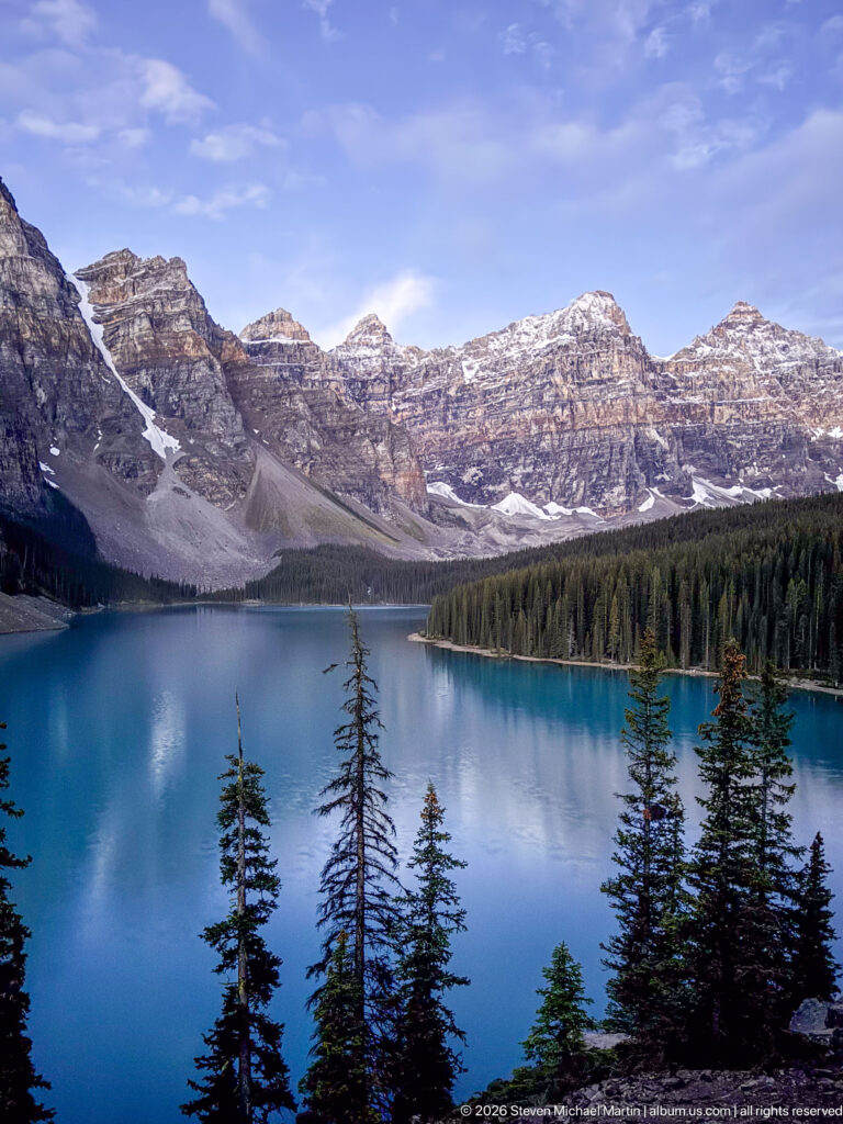 Blue Moraine Lake below a forested shoreline and broad layered mountain cliffs, with foreground conifers and patches of snow.