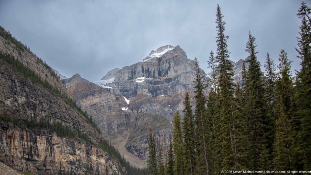 Layered gray mountain cliffs with patches of snow rising behind tall evergreen trees under cloudy sky.