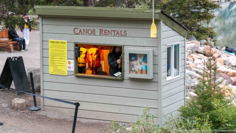 Small gray canoe-rental kiosk with service window, yellow information board, and Canoe Rentals sign.