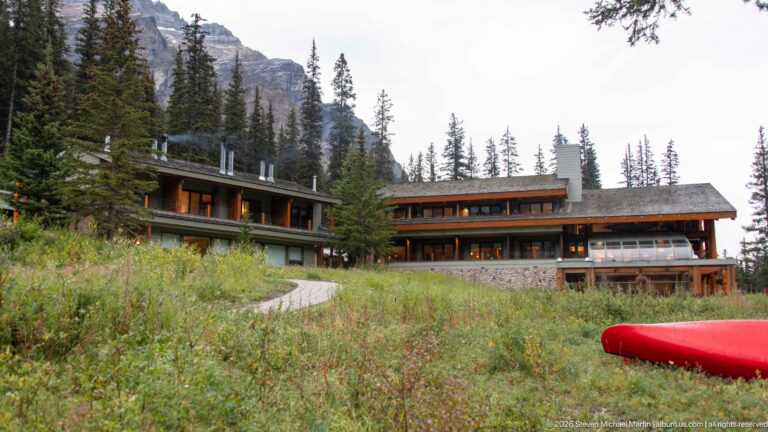 Low timber lodge buildings in a mountain clearing with wild vegetation and a red canoe in the foreground.