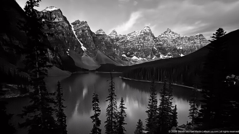 Black-and-white view of Moraine Lake with silhouetted conifers, reflective water, jagged snowy peaks, and heavy clouds.