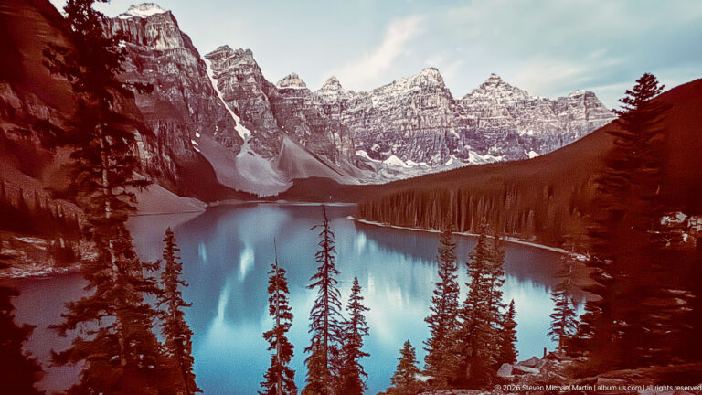 Warm red-toned mountain slopes surround bright cyan Moraine Lake, with conifers in the foreground and jagged peaks beyond.
