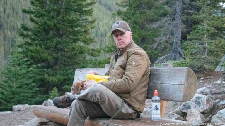 Visitor in brown outdoor clothing seated on a log bench with food and a drink amid conifers and rocks.