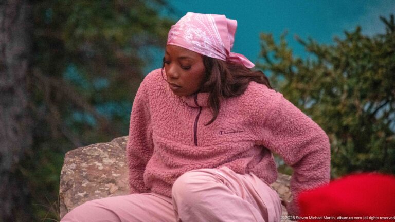 Visitor wearing pink outdoor clothing and a headscarf seated on a boulder, with blurred conifers behind.