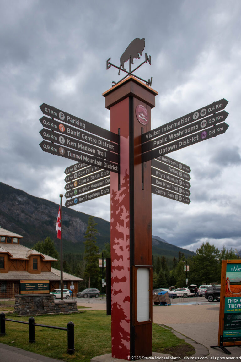 Red multi-directional Banff wayfinding sign topped by an animal weather vane, with mountains and low buildings behind.