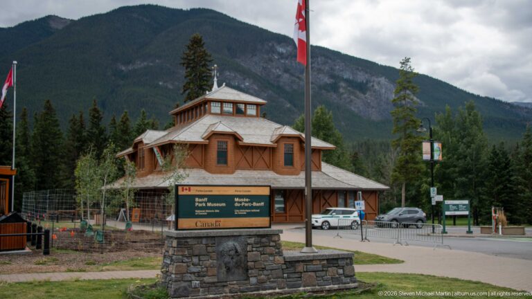 Banff Park Museum behind a stone-based bilingual site sign, with a Canadian flag and mountain slope in the background.